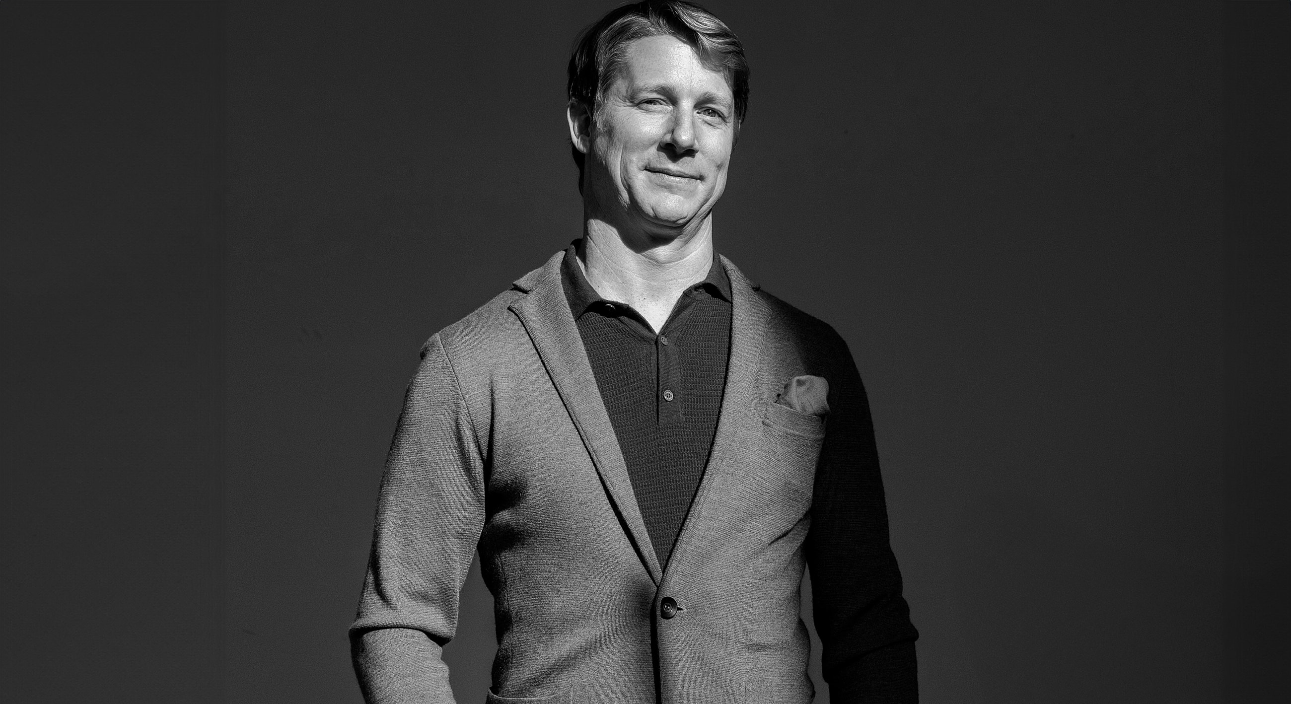 Man in suit smiling against dark background.